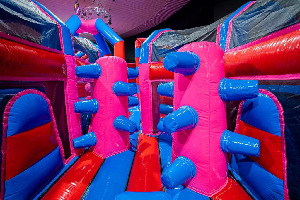 Obstáculo hinchable con postes verticales y brazos de empuje azules en un parque hinchable colorido